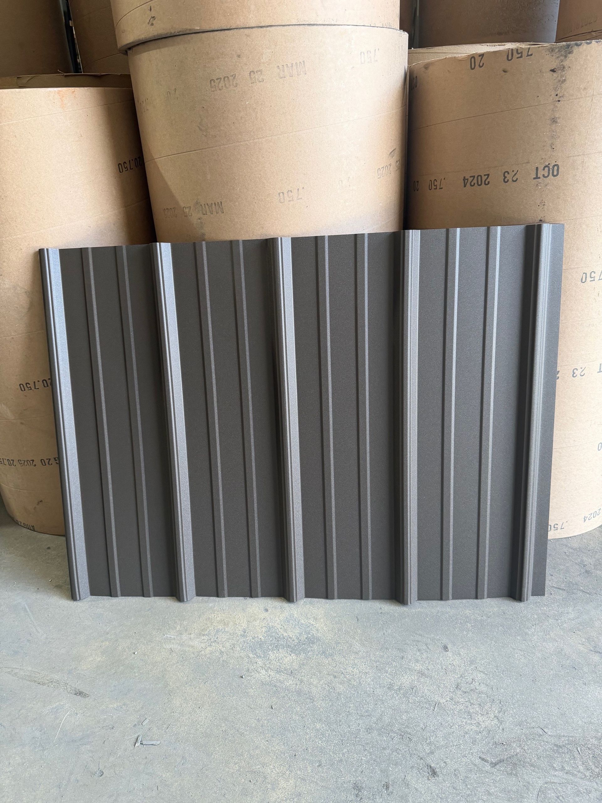 A dark brown, rectangular metal roofing panel with vertical ribbing, standing against a background of paper rolls.