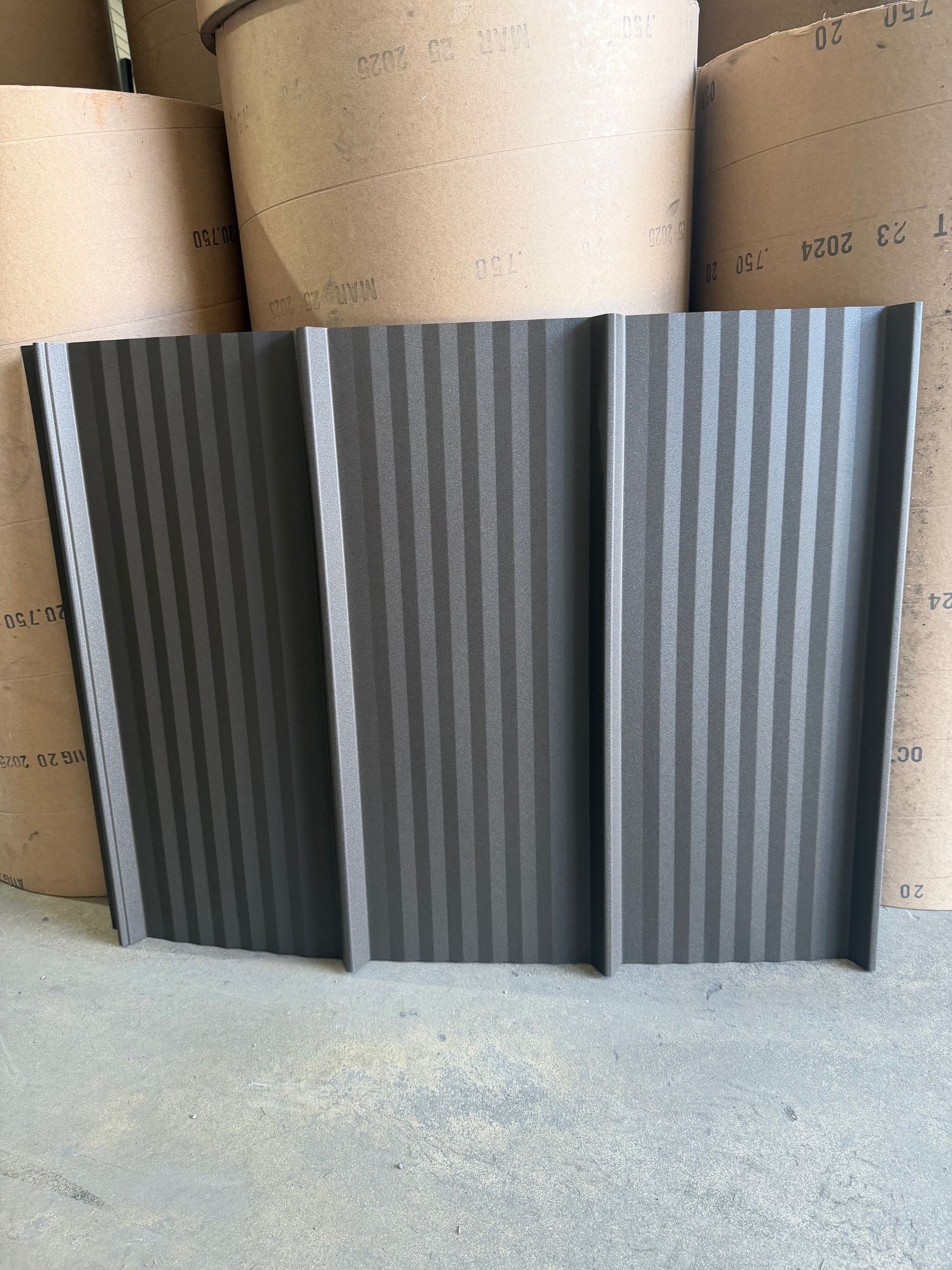 A dark grey, corrugated metal roofing panel standing upright in front of several large, stacked paper rolls.