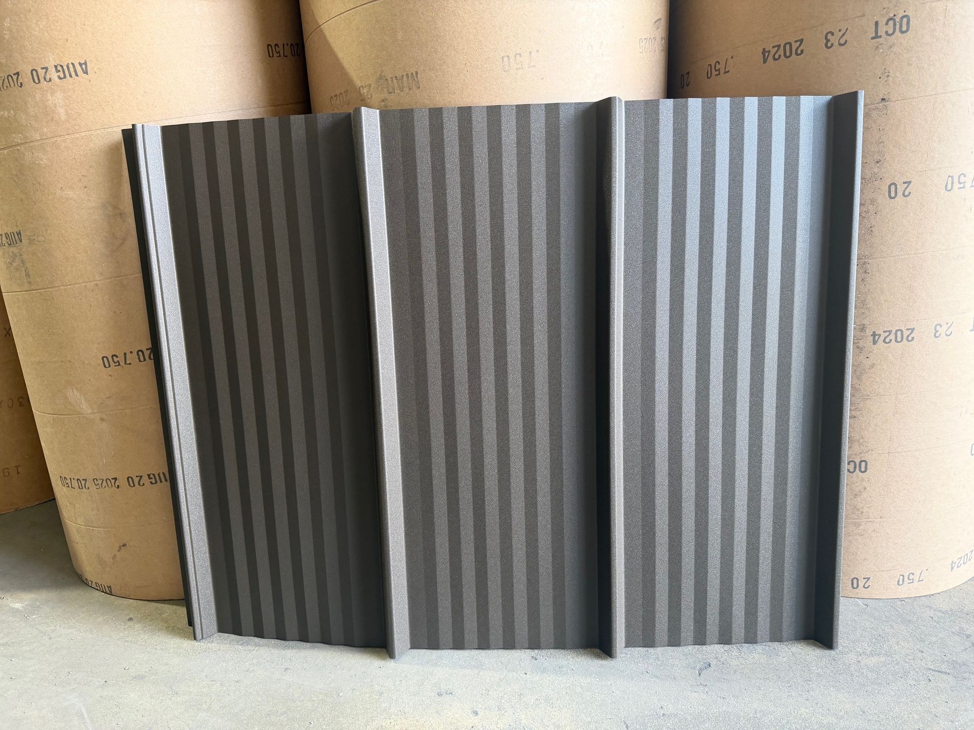 A dark gray, vertically corrugated metal panel standing against rolls of industrial paper.