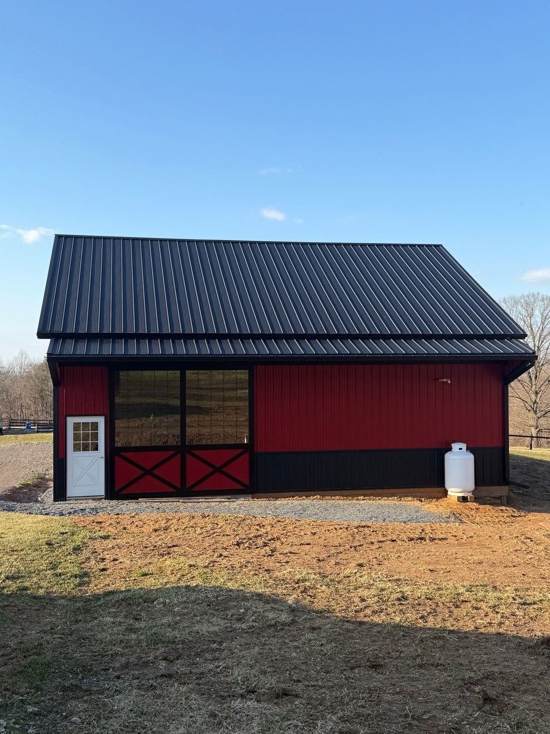 A red and black metal barn with a white door and a large glass window stands on a gravel lot under a clear blue sky.