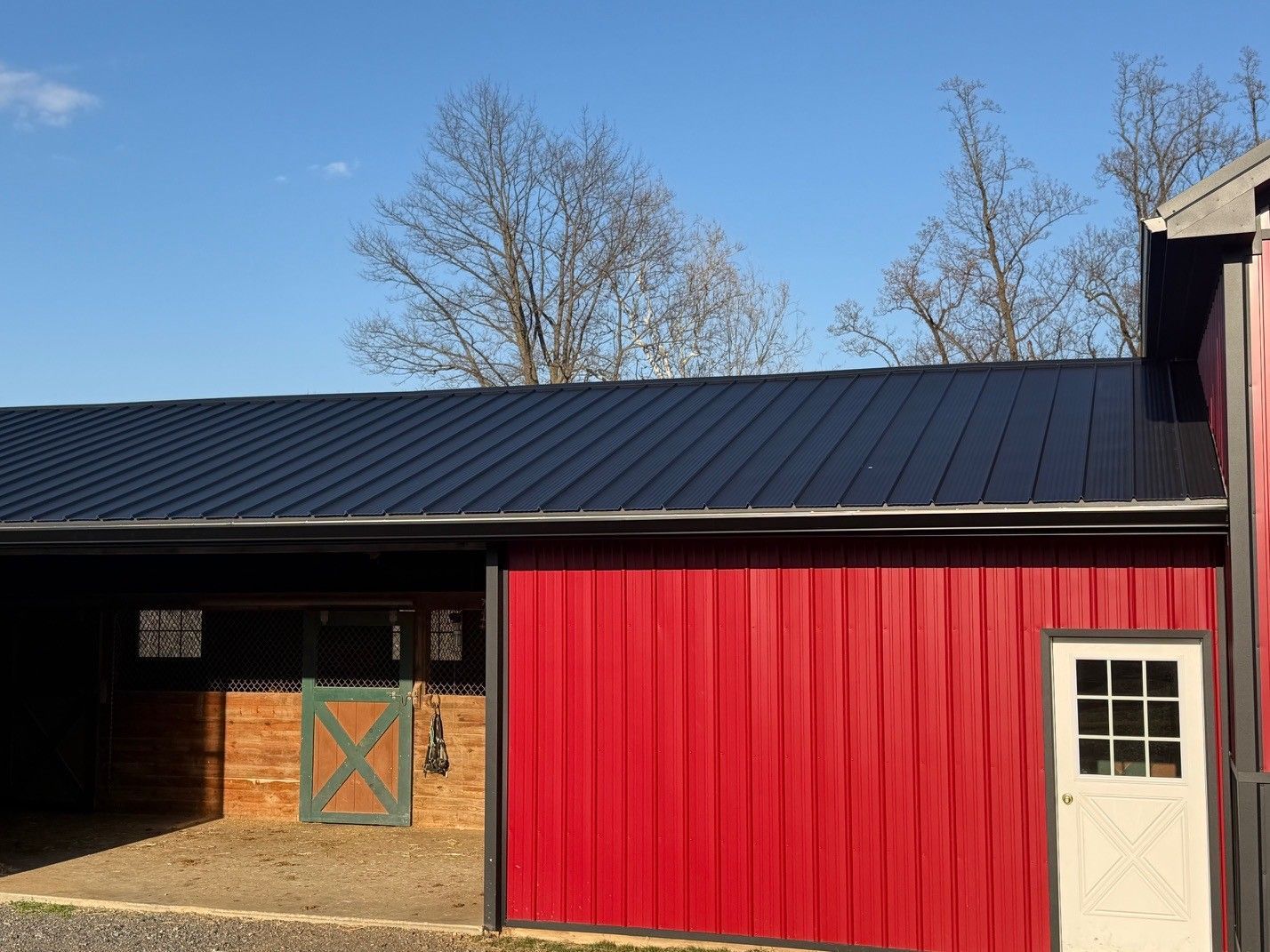 A red barn with a dark metal roof, featuring a horse stall, a white door, and trees against a blue sky.