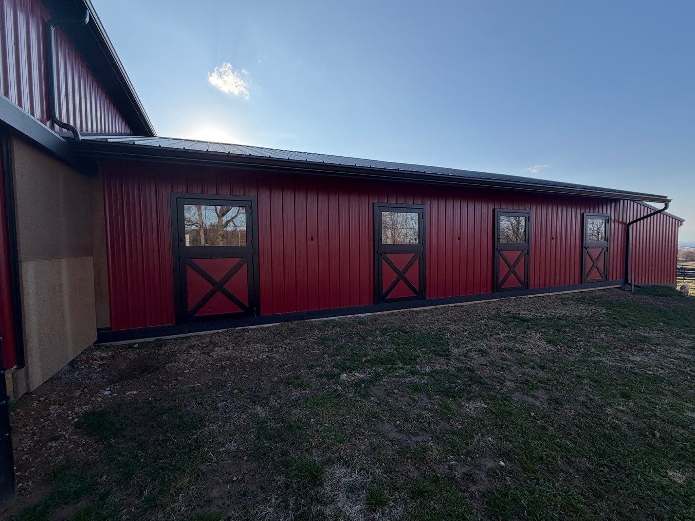 A bright red barn with a metal roof and a row of four black-trimmed stall doors, set against a clear blue sky.
