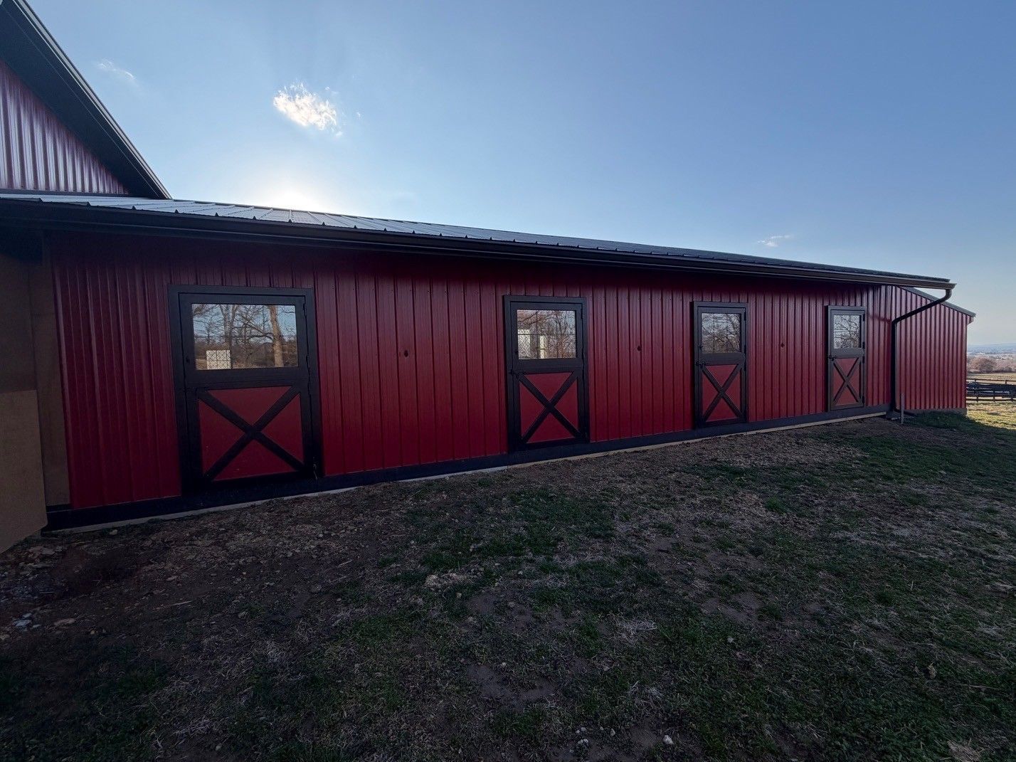 Red barn exterior with four black-framed doors, featuring cross-buck detailing, on a grassy hillside under a blue sky.