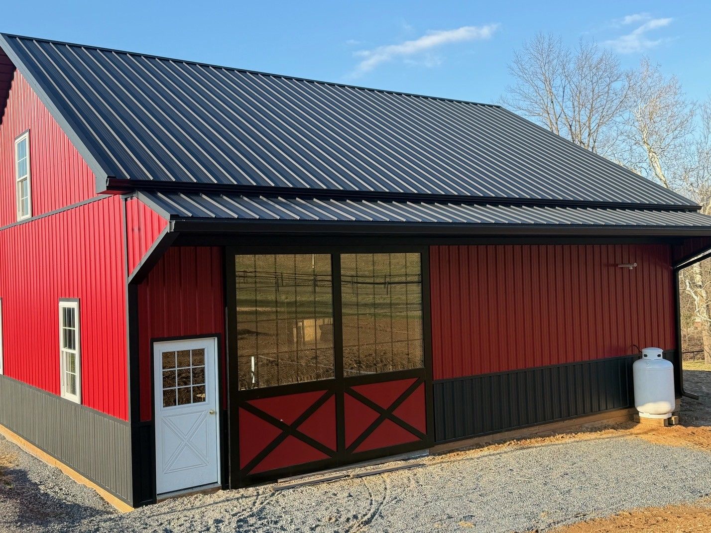 Red and black metal barn with a white door, large sliding window, and metal roof, situated on a gravel lot under blue sky.