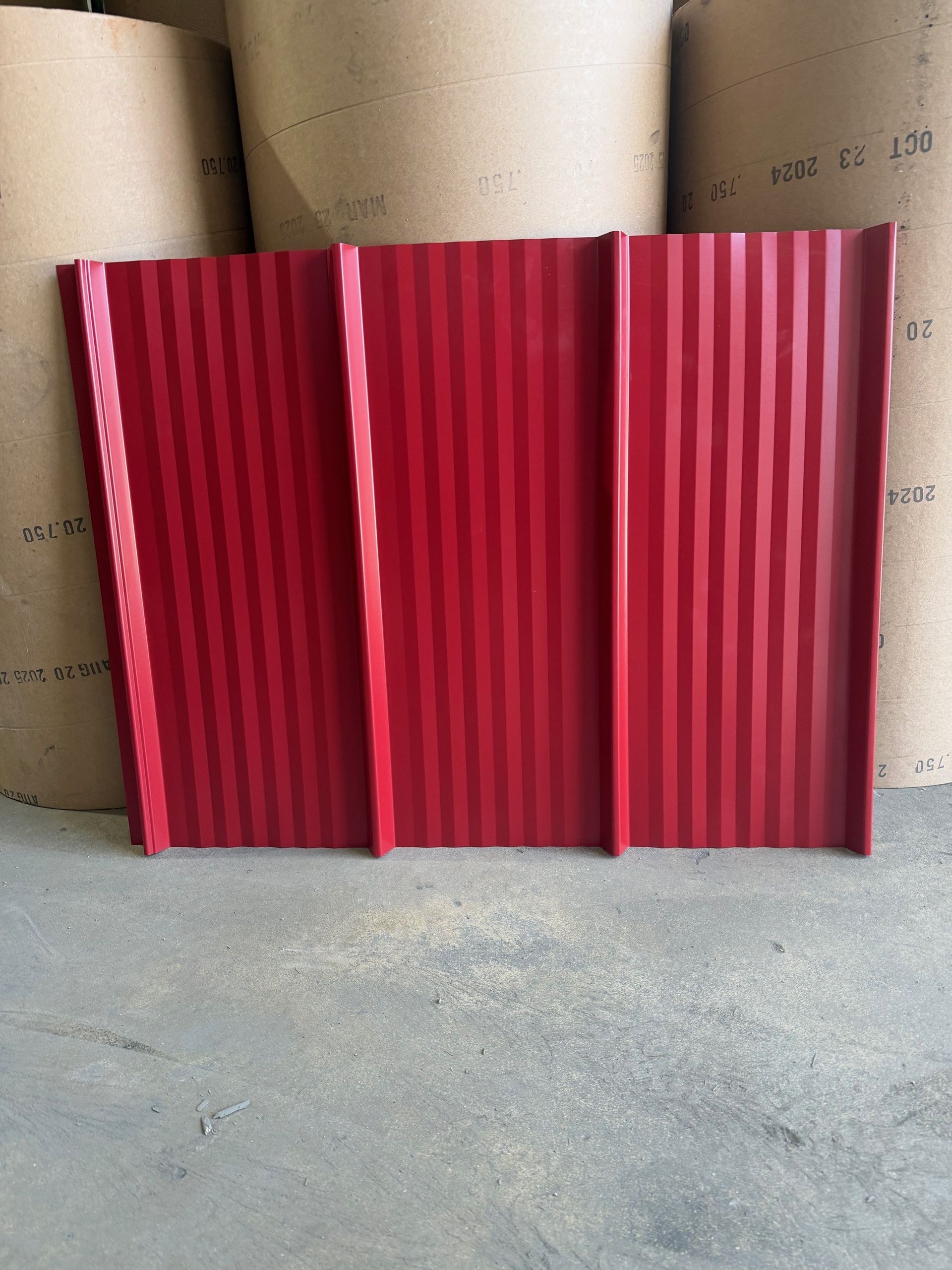 A red, corrugated metal roofing sheet standing upright in front of large rolls of paper or insulation materials.