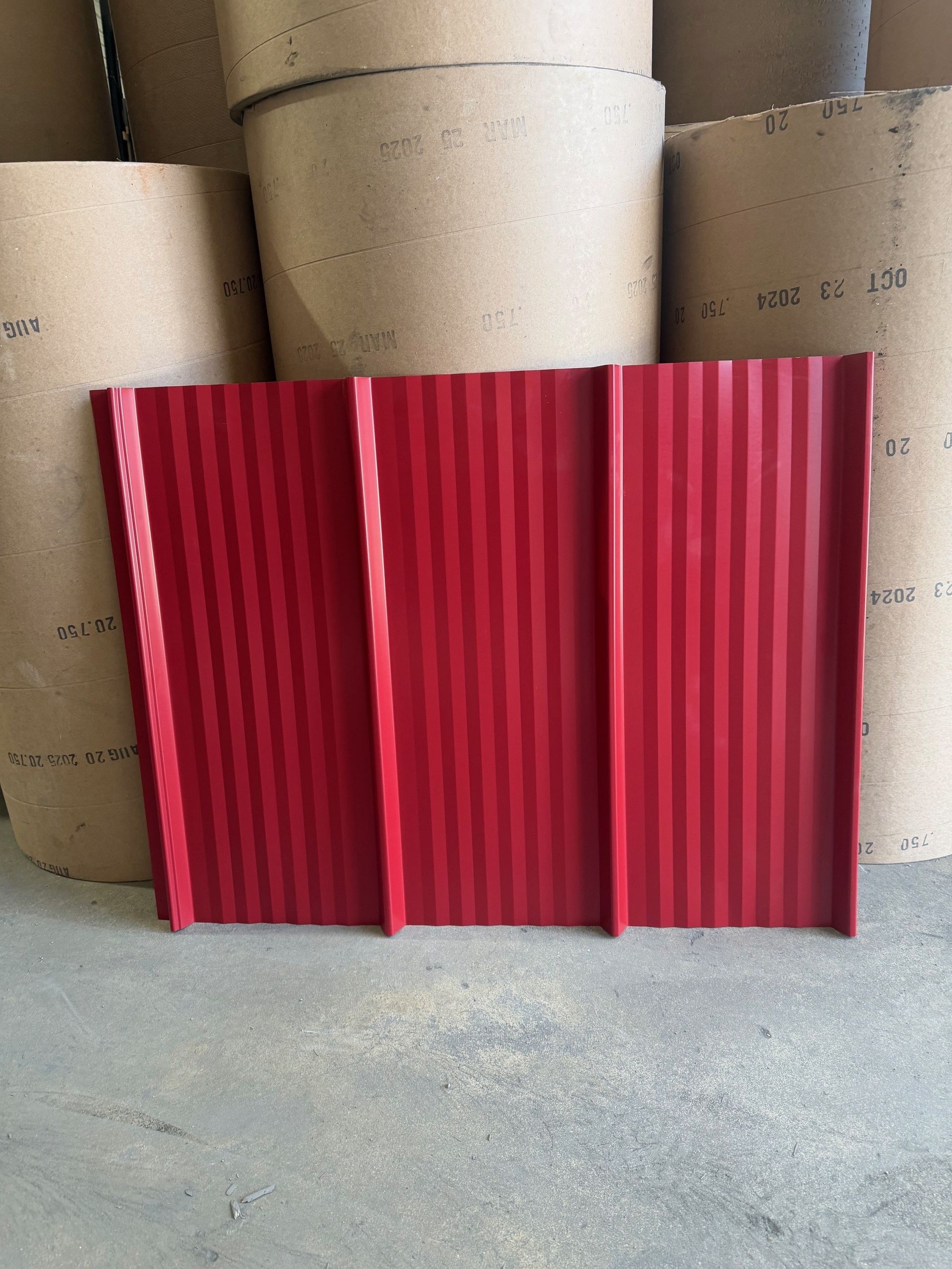 A red corrugated metal roofing panel standing upright against stacked paper rolls in a warehouse setting.