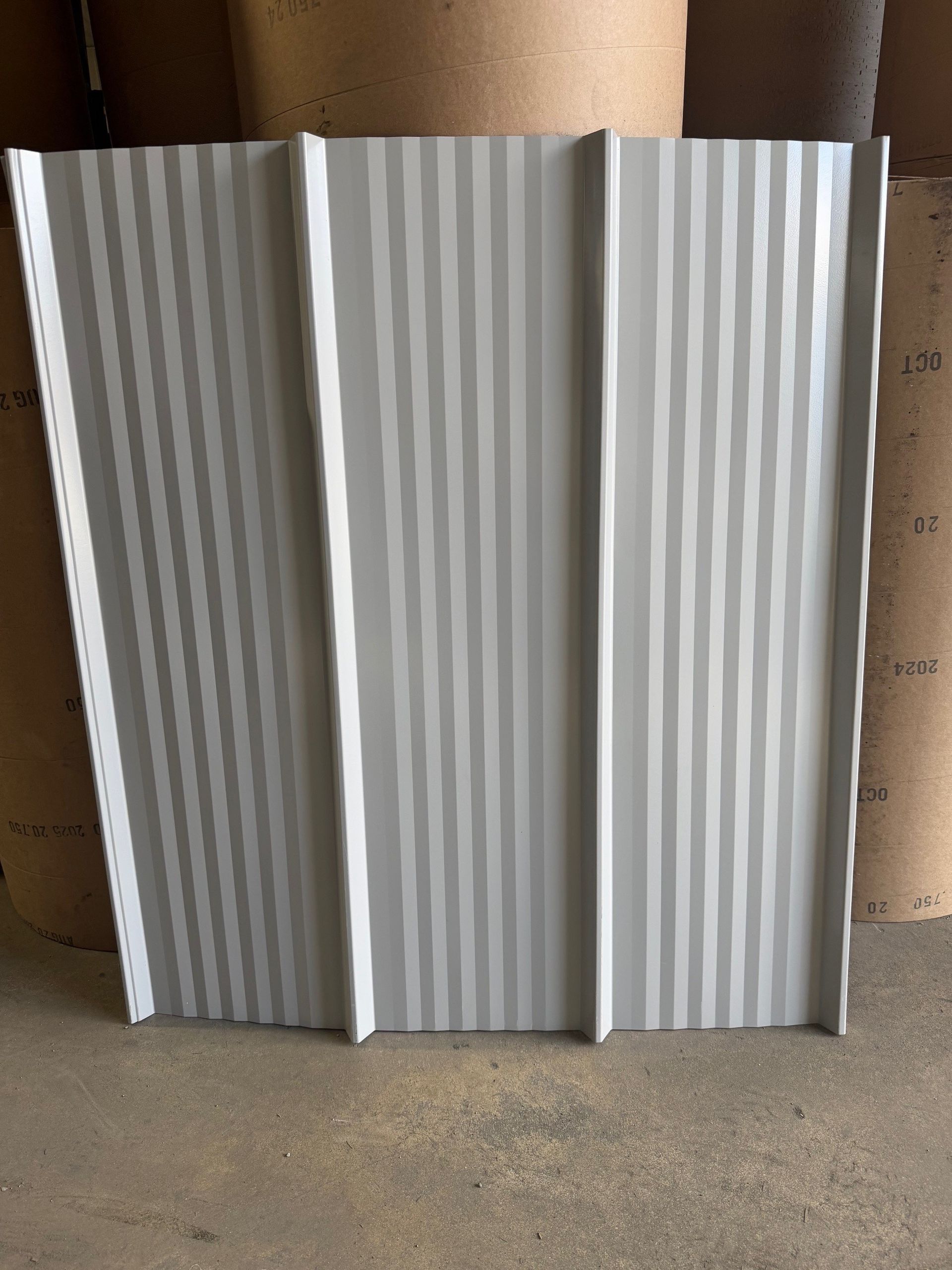 A light gray metal roofing panel with three vertical ridges, standing upright against a background of stacked paper rolls.