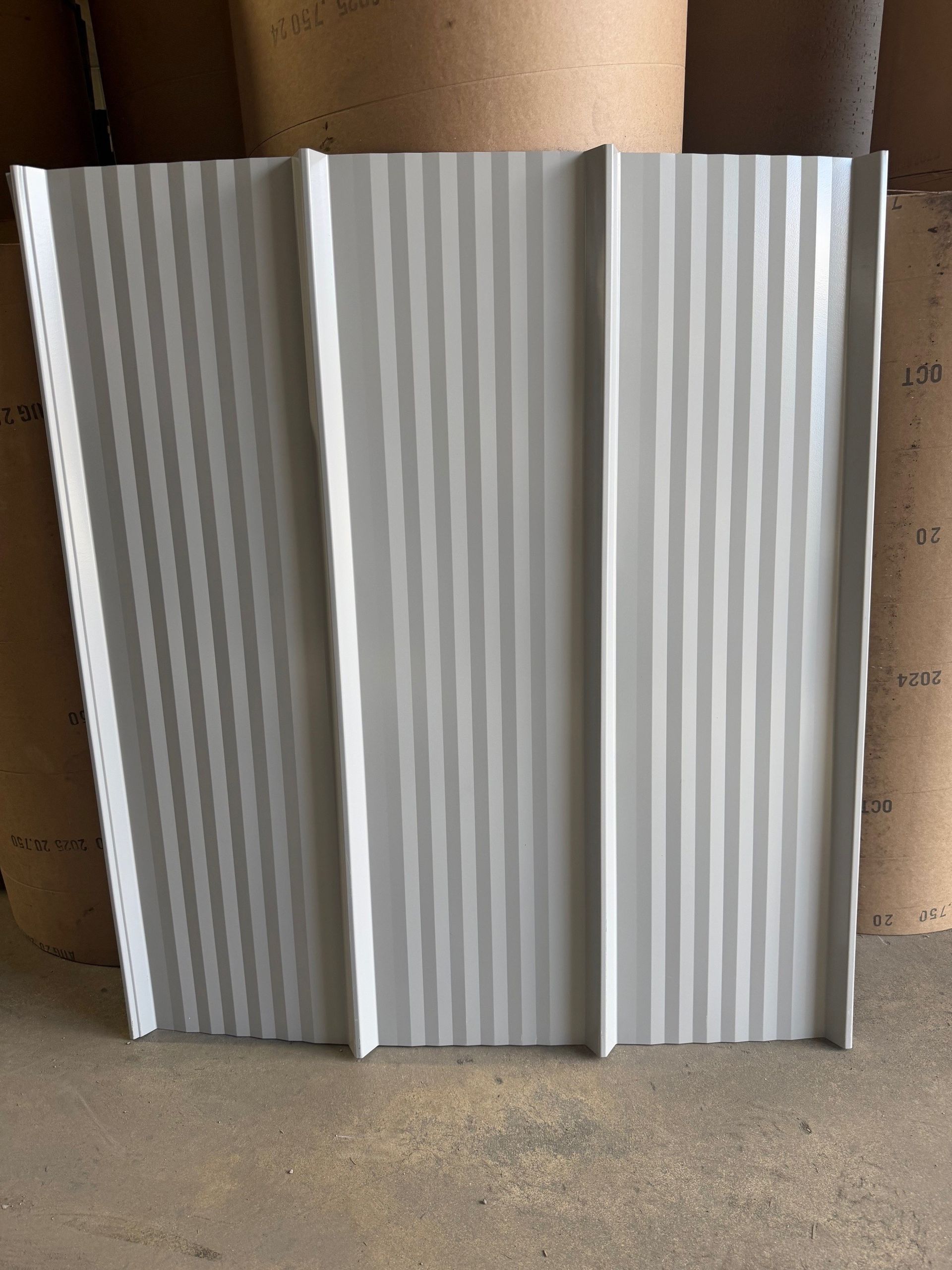 A rectangular light grey metal roofing or siding panel with raised vertical ribs, standing upright on a concrete floor.