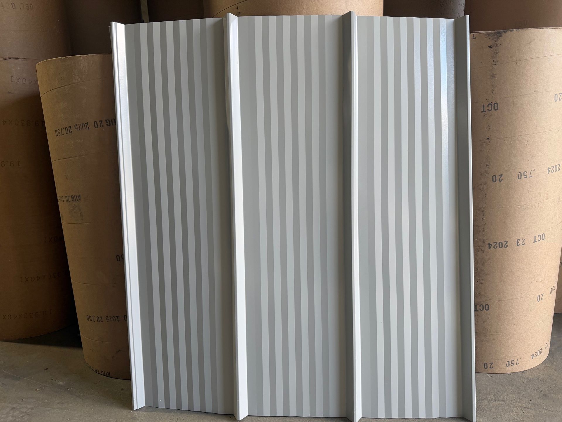 A light-colored, ribbed metal roofing or siding panel standing vertically in front of several large rolls of paper.