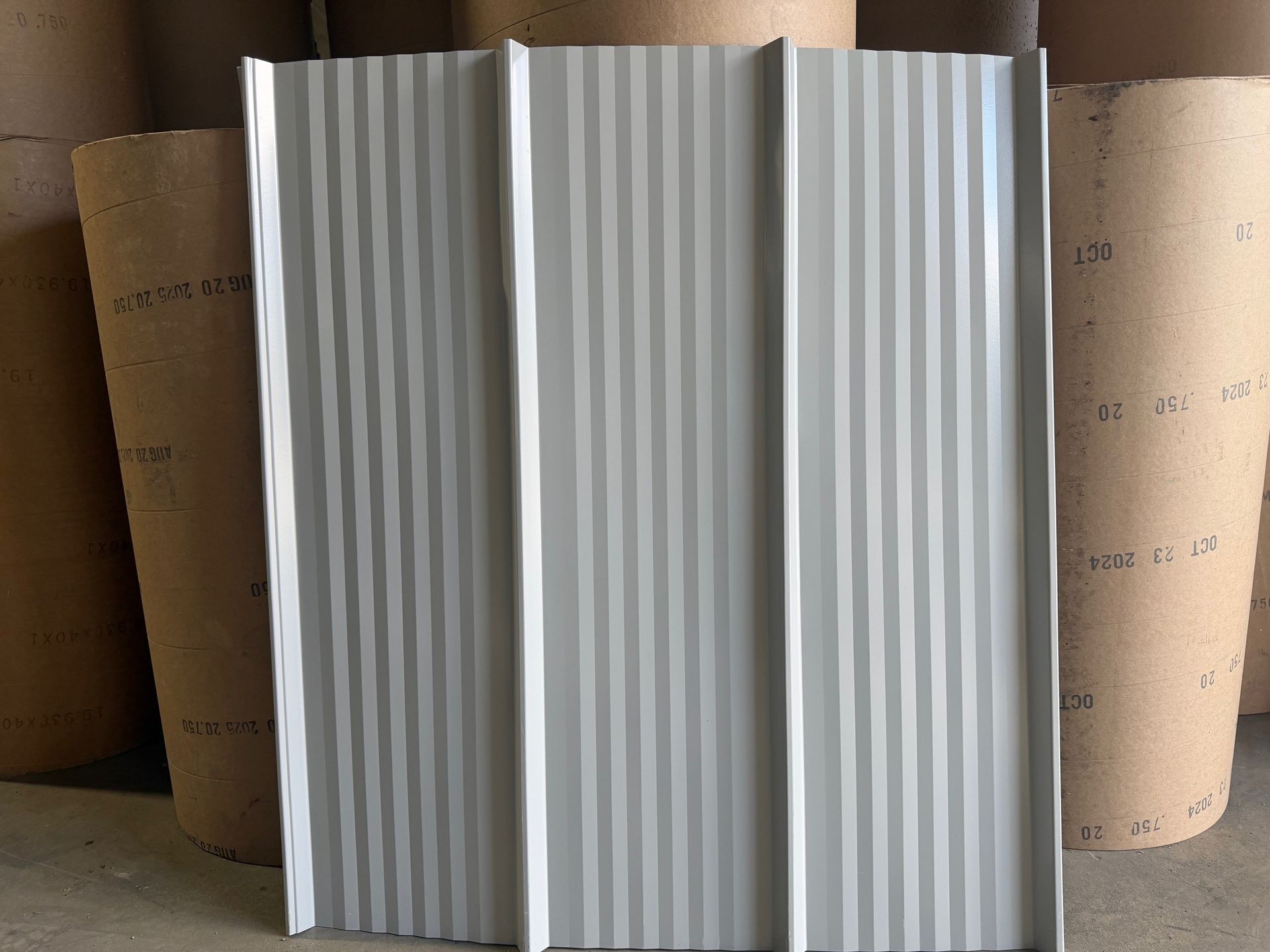 A light gray metal roofing panel with vertical ribbing, standing in front of several large rolls of brown paper.