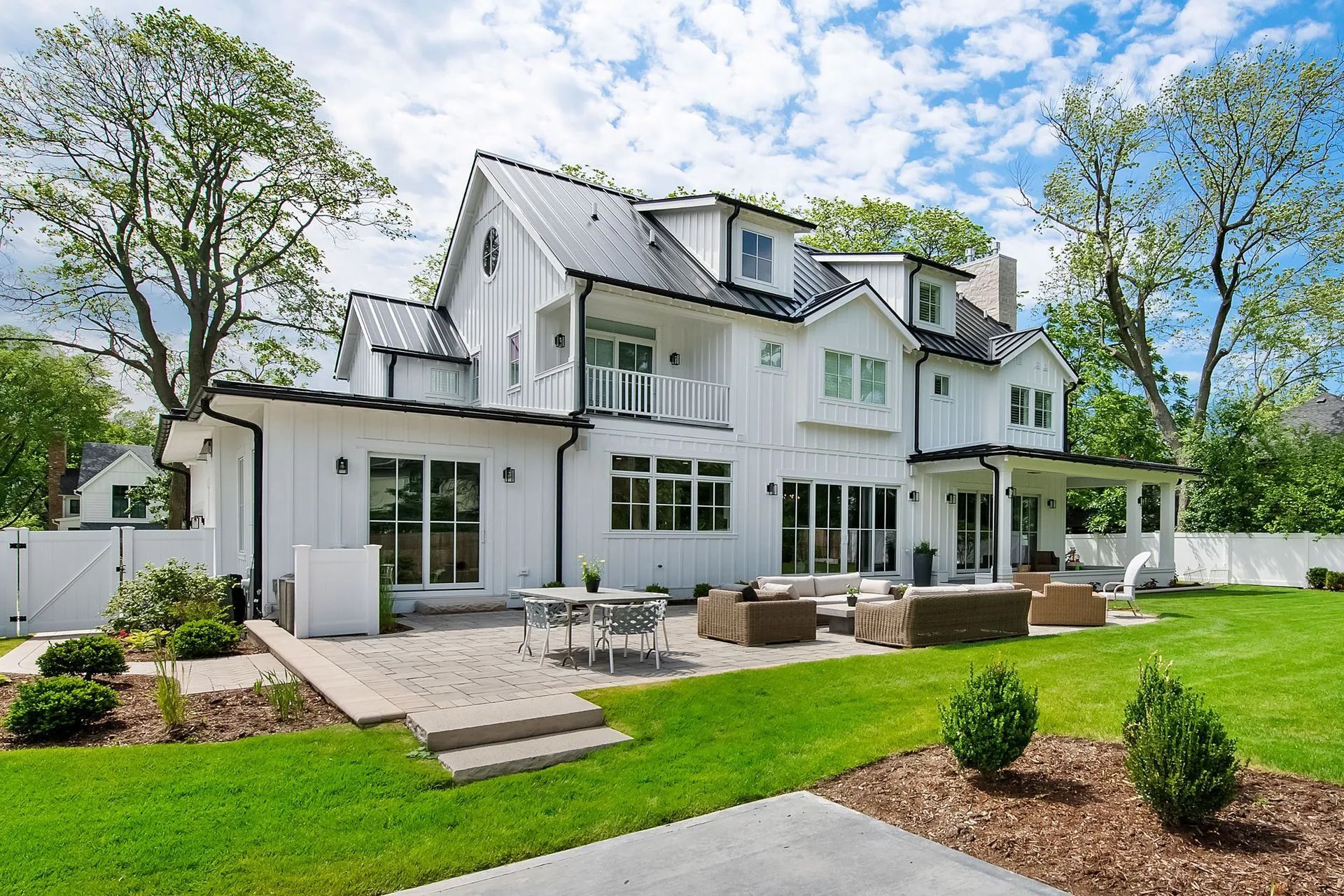 A large, white, multi-story modern farmhouse with a stone patio, outdoor seating, and a lush green lawn under a blue sky.