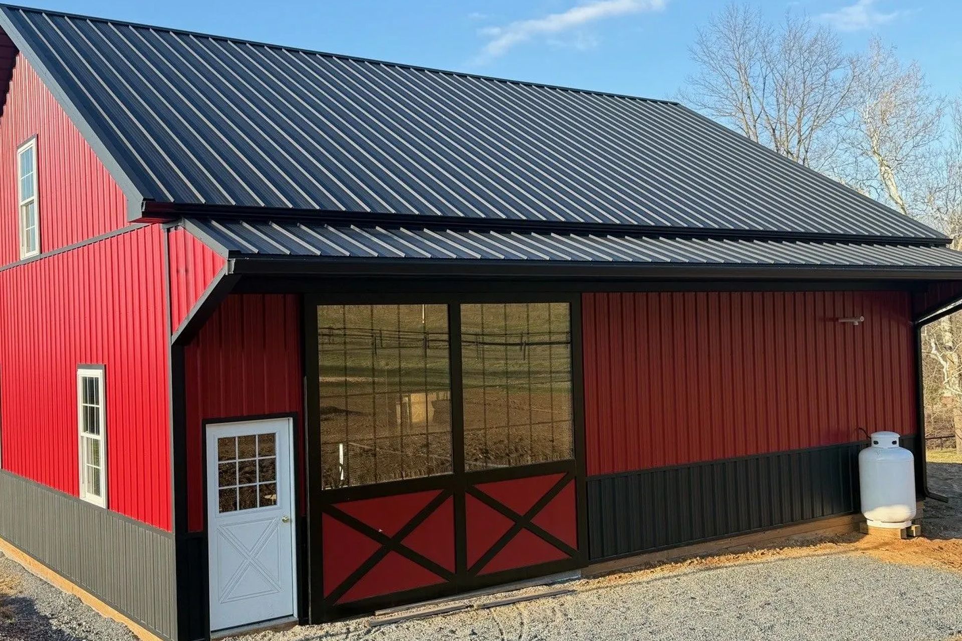 A red and black pole barn building with a metal roof and large glass sliding doors, set on a gravel surface.