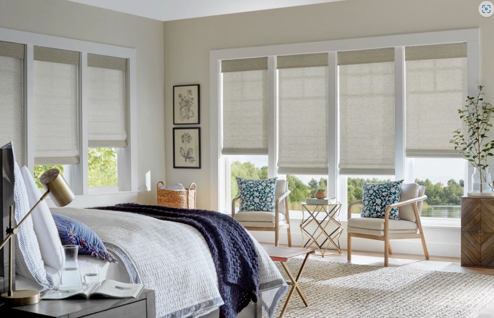 A bedroom with a bed , chairs , and a window with roman shades.