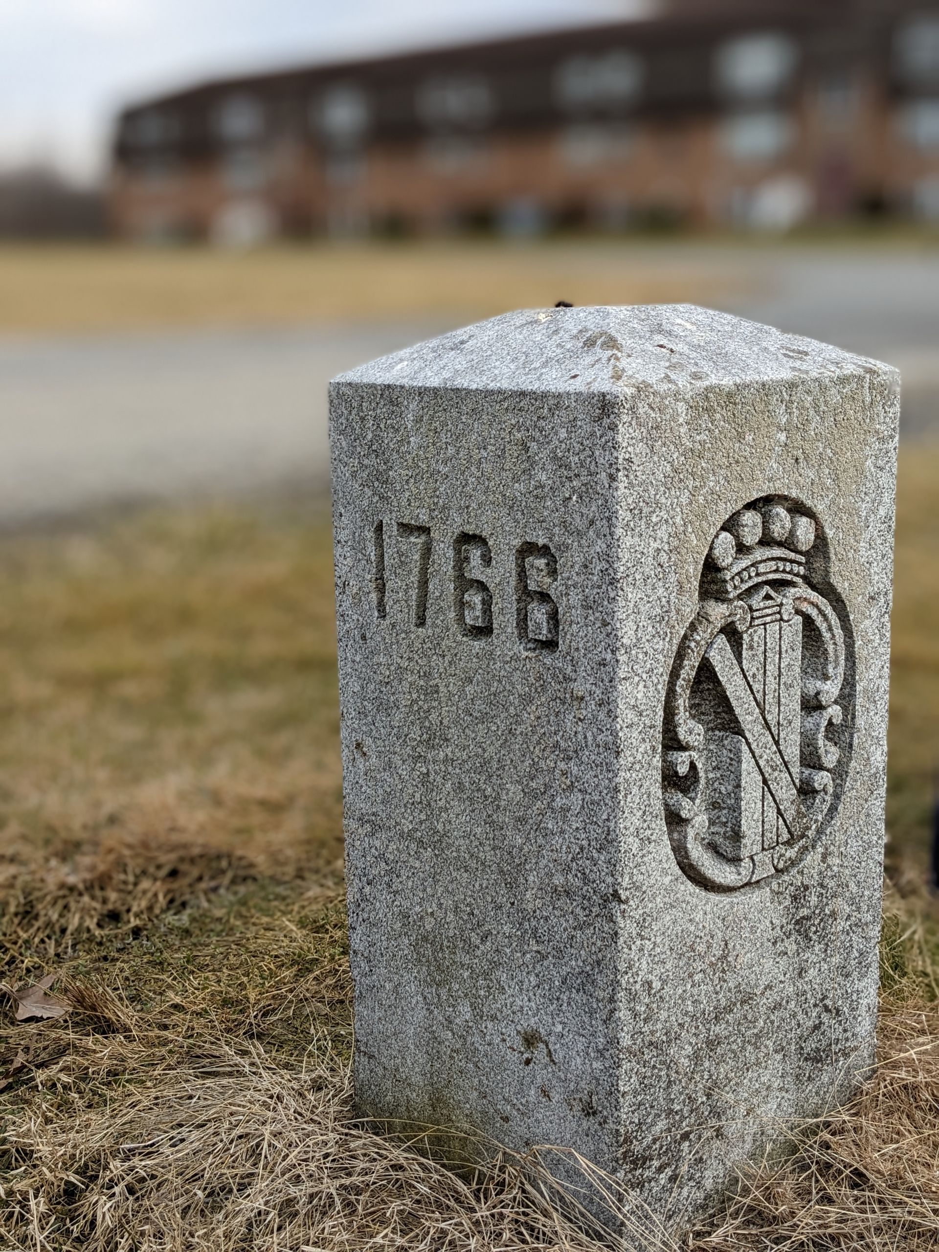 Granite marker from 1766, with engraved text and emblem, set in grass. Building in the soft focus background.