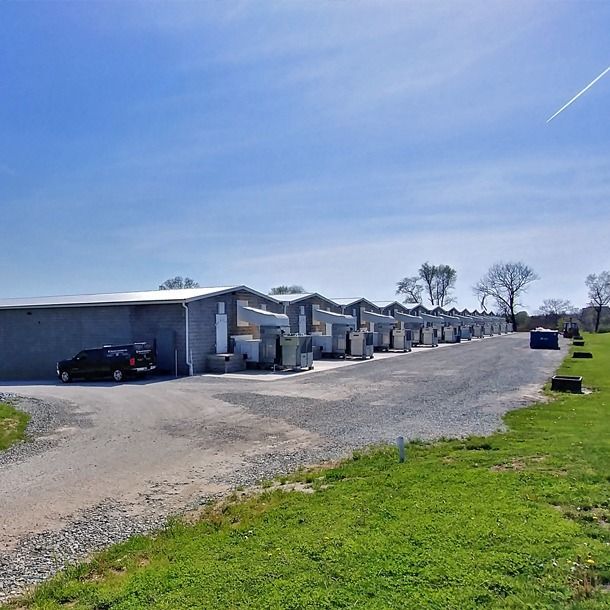 Row of gray storage units with gravel driveway, blue sky, and green grass.