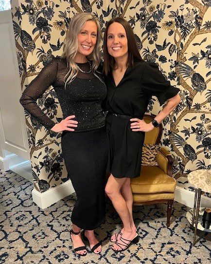 Two women in black outfits pose by floral wallpaper and a patterned rug.
