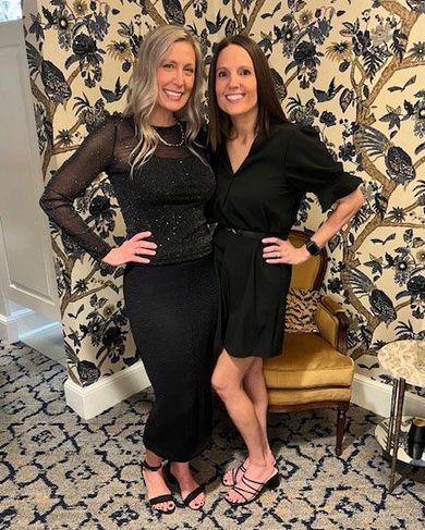 Two women in black outfits pose by floral wallpaper and a patterned rug.