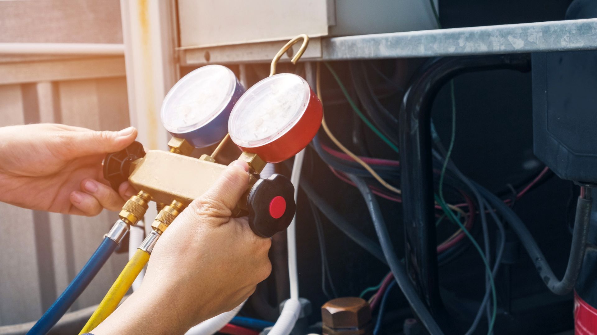 Hands using gauges to service an HVAC unit; hoses, wires visible.