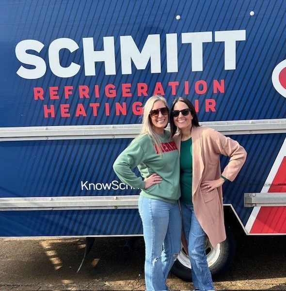 Two women pose in front of a blue Schmitt Refrigeration truck. One wears a green sweatshirt, the other a tan coat.