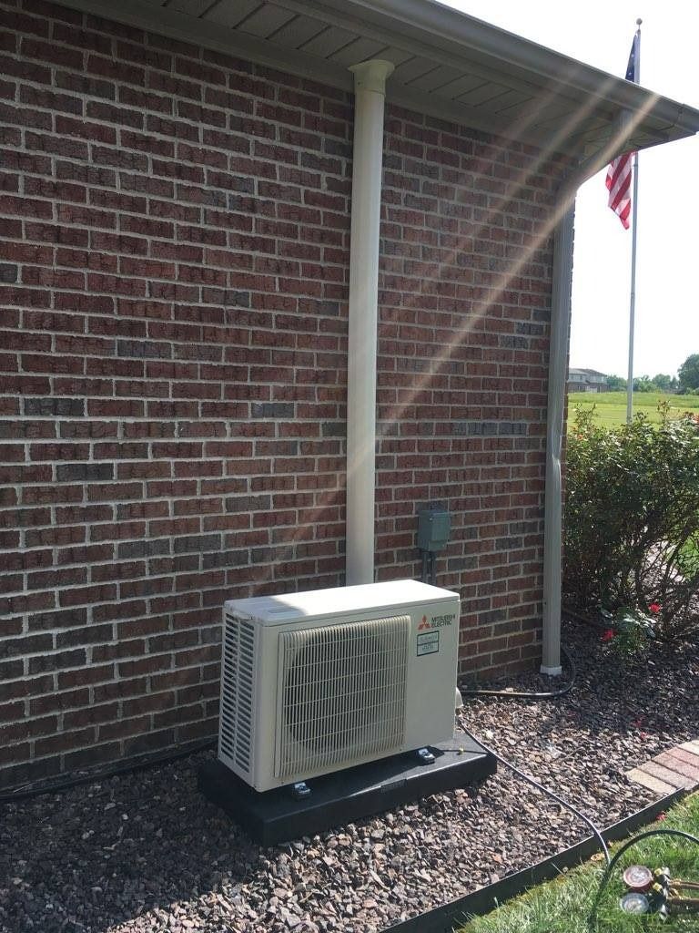 Exterior air conditioning unit next to a brick wall and a white pipe.
