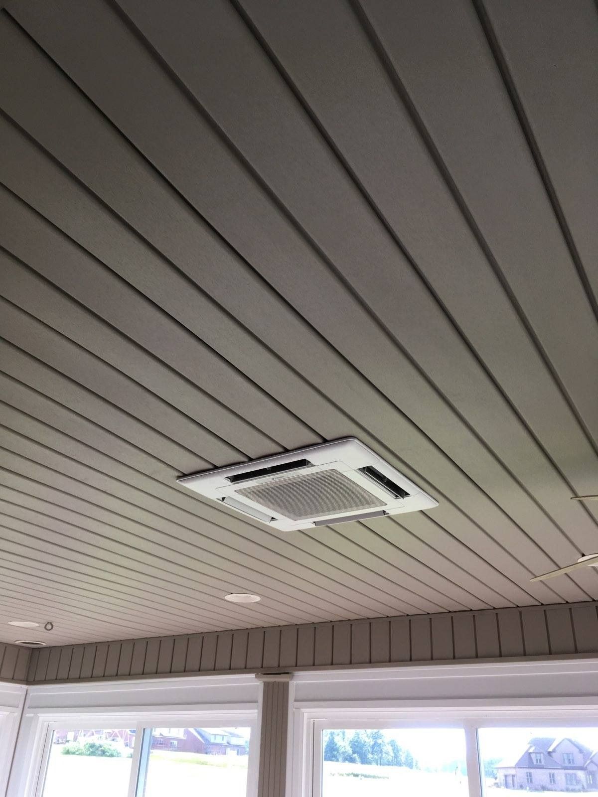 Ceiling-mounted air conditioner in a room with a slatted gray ceiling and windows below.