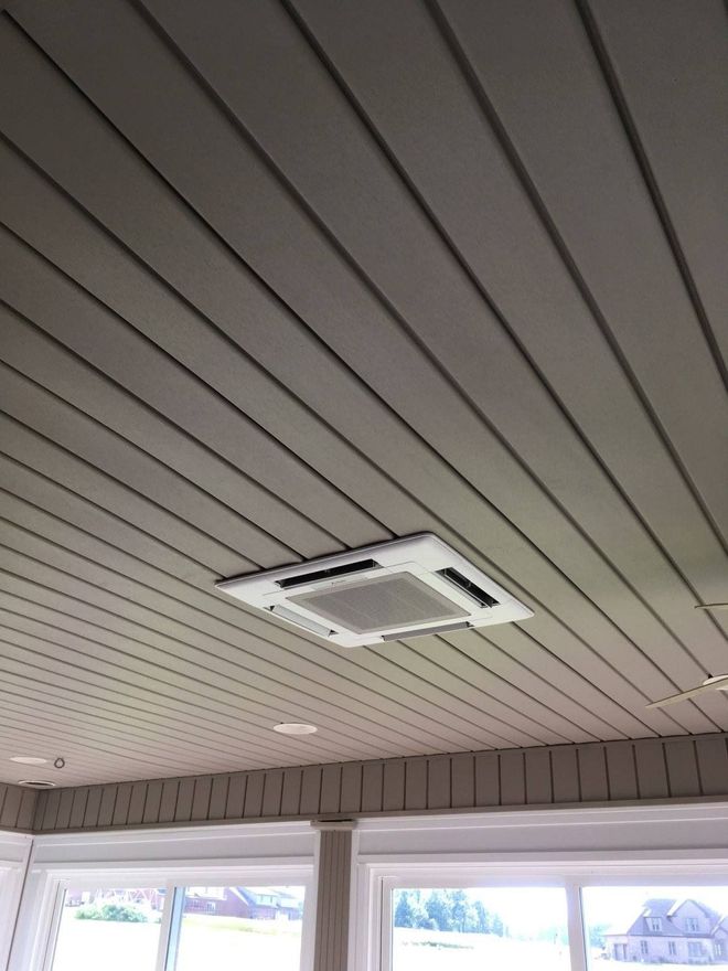 White ceiling-mounted HVAC unit against gray, grooved ceiling panels in a room with windows and trim.