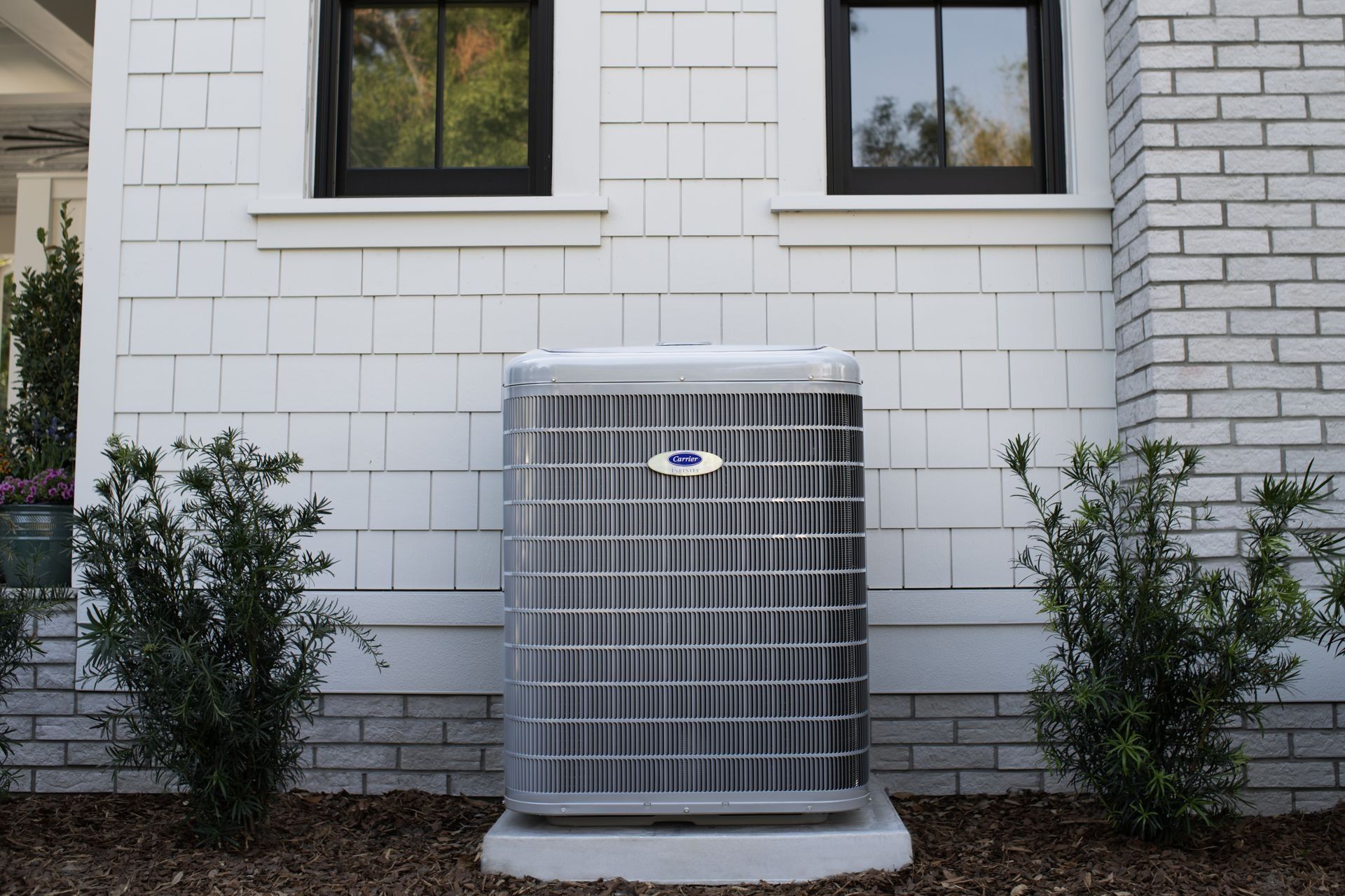 Air conditioning unit outside a white shingle-sided building with two windows.