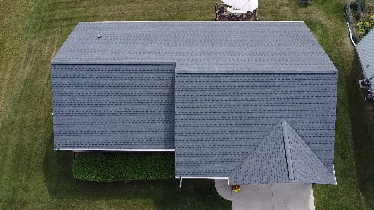 An aerial view of a gray shingled residential roof with an L-shaped design and a small gabled section above a driveway.