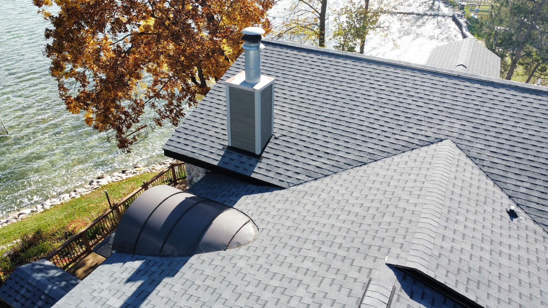 An elevated view of a gray shingled roof featuring a metal chimney, a rounded roof vent, and a lake in the background.
