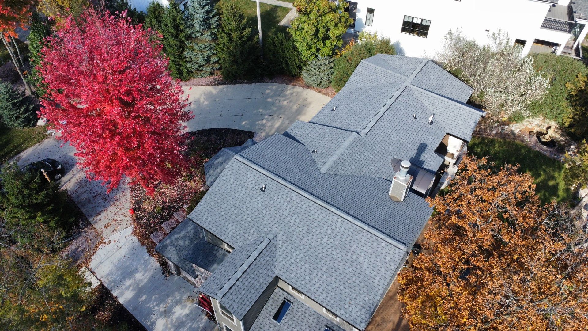 An aerial view of a house roof surrounded by autumn trees, including one with bright red foliage.