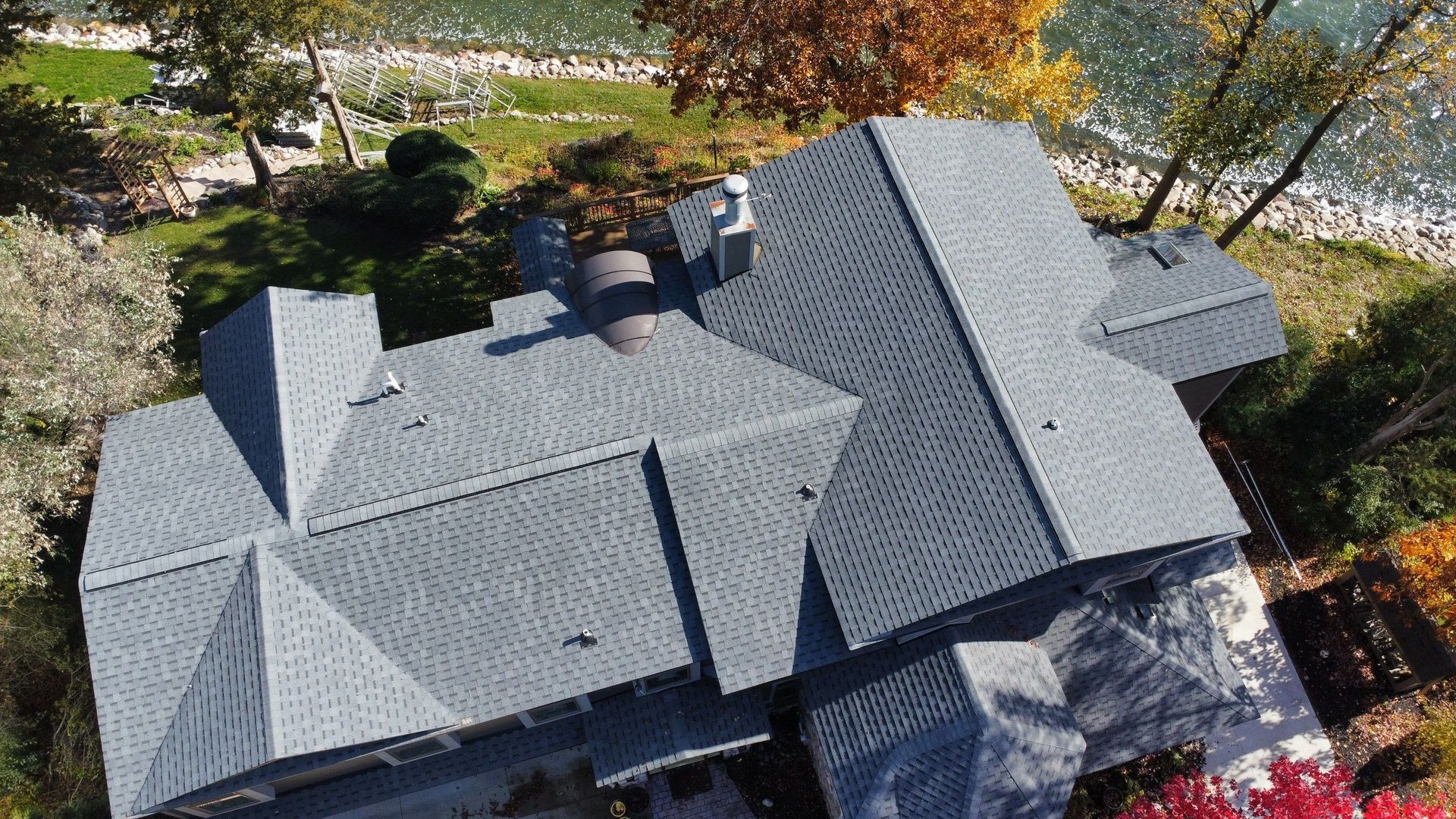 An aerial view of a large house with a grey, multi-angled shingled roof, surrounded by trees near a body of water.