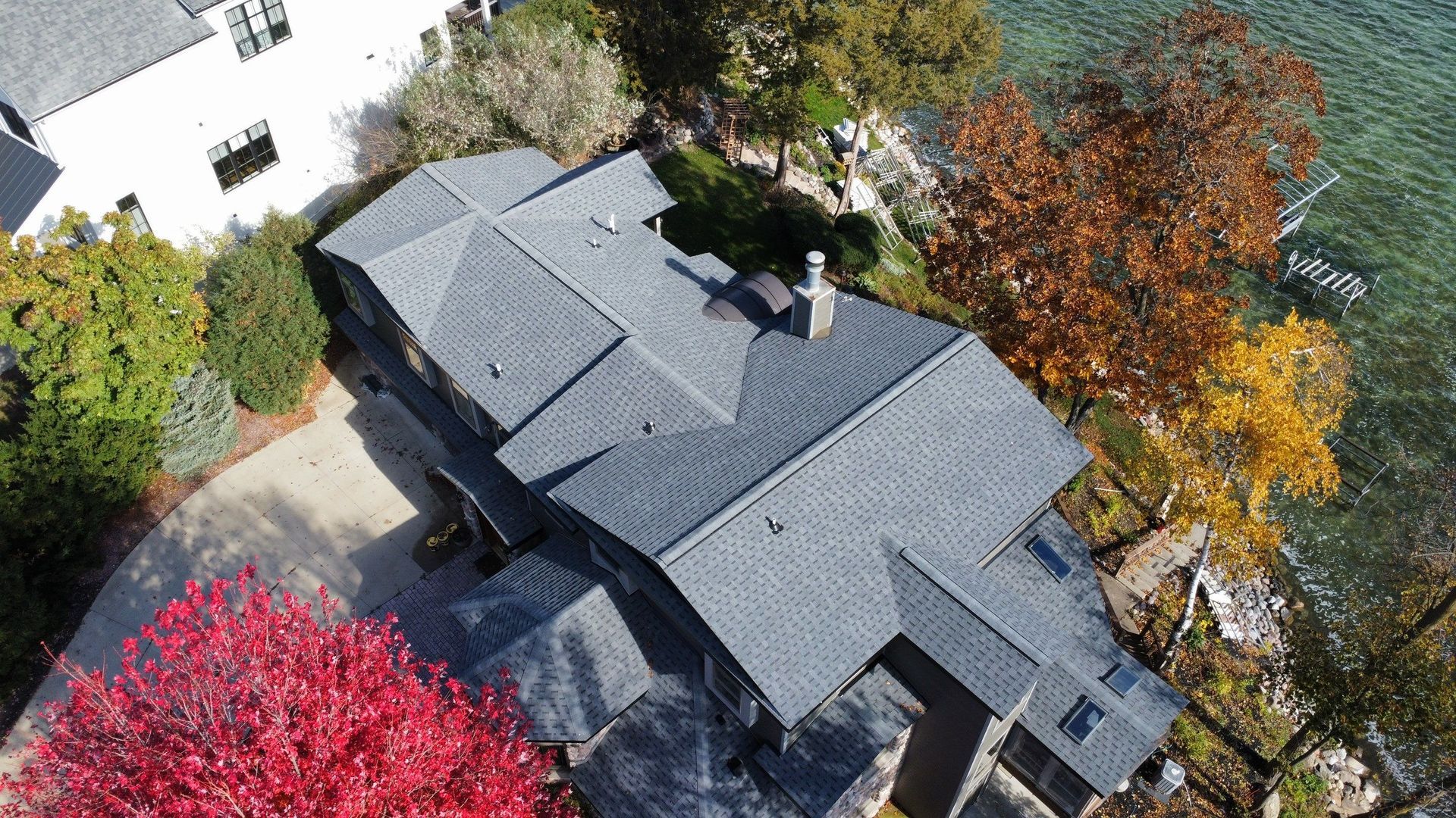 An aerial view of a dark shingled house nestled among autumn trees by a lake, with a bright red tree in the foreground.