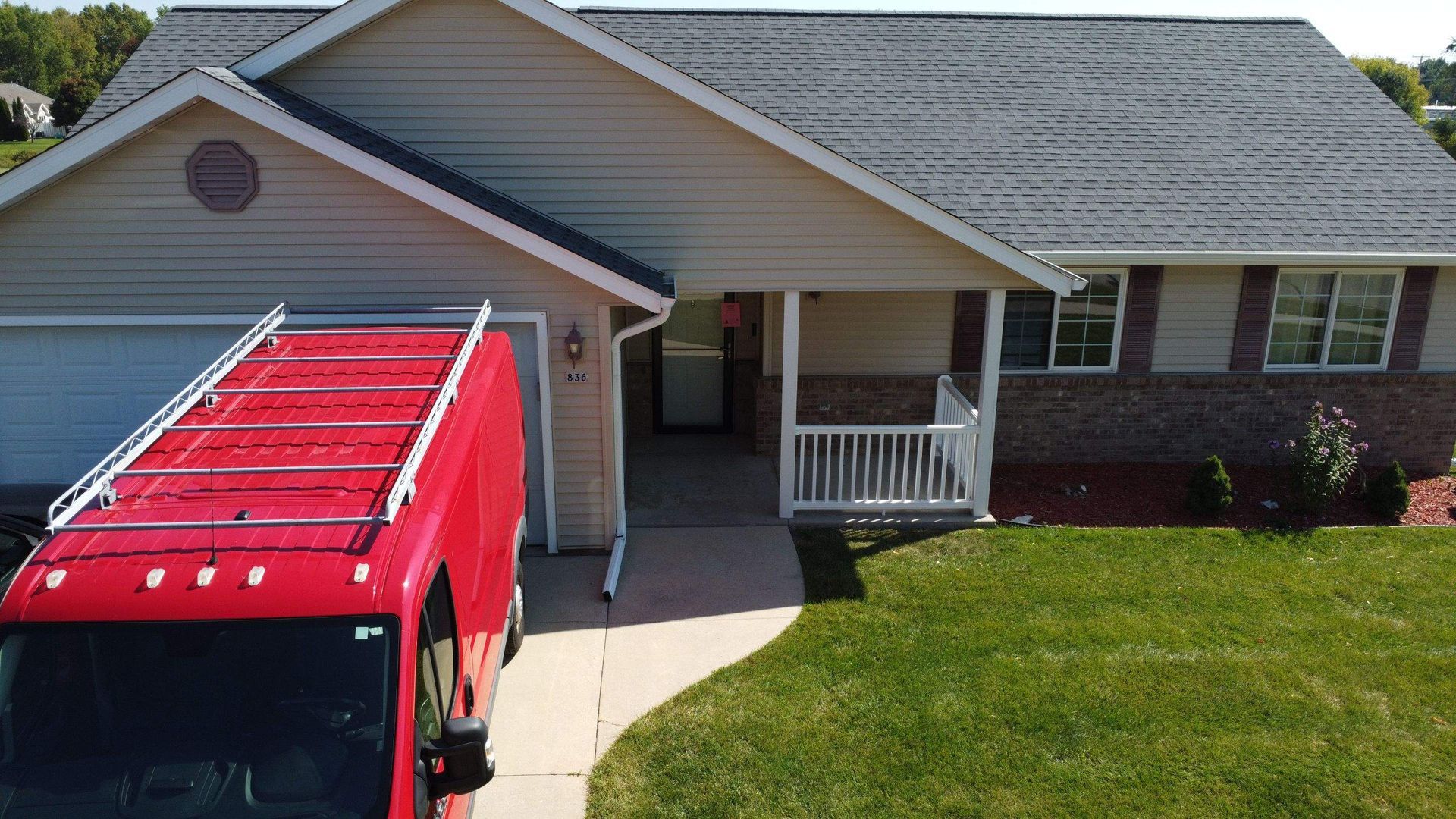A bright red utility van with a roof rack parked in the driveway of a tan suburban home with a grassy lawn.