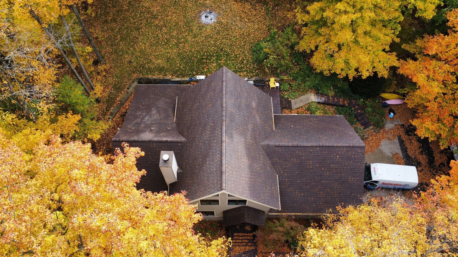Aerial view of a house with a shingled roof surrounded by dense autumn trees with bright yellow and orange leaves.