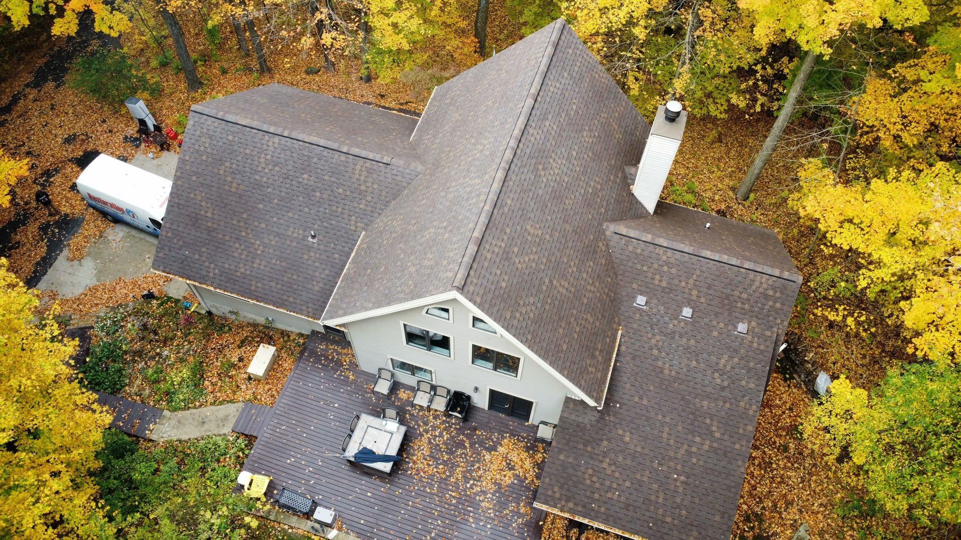 Aerial view of a house with a shingled roof, surrounded by trees with yellow autumn leaves, and a deck with furniture.