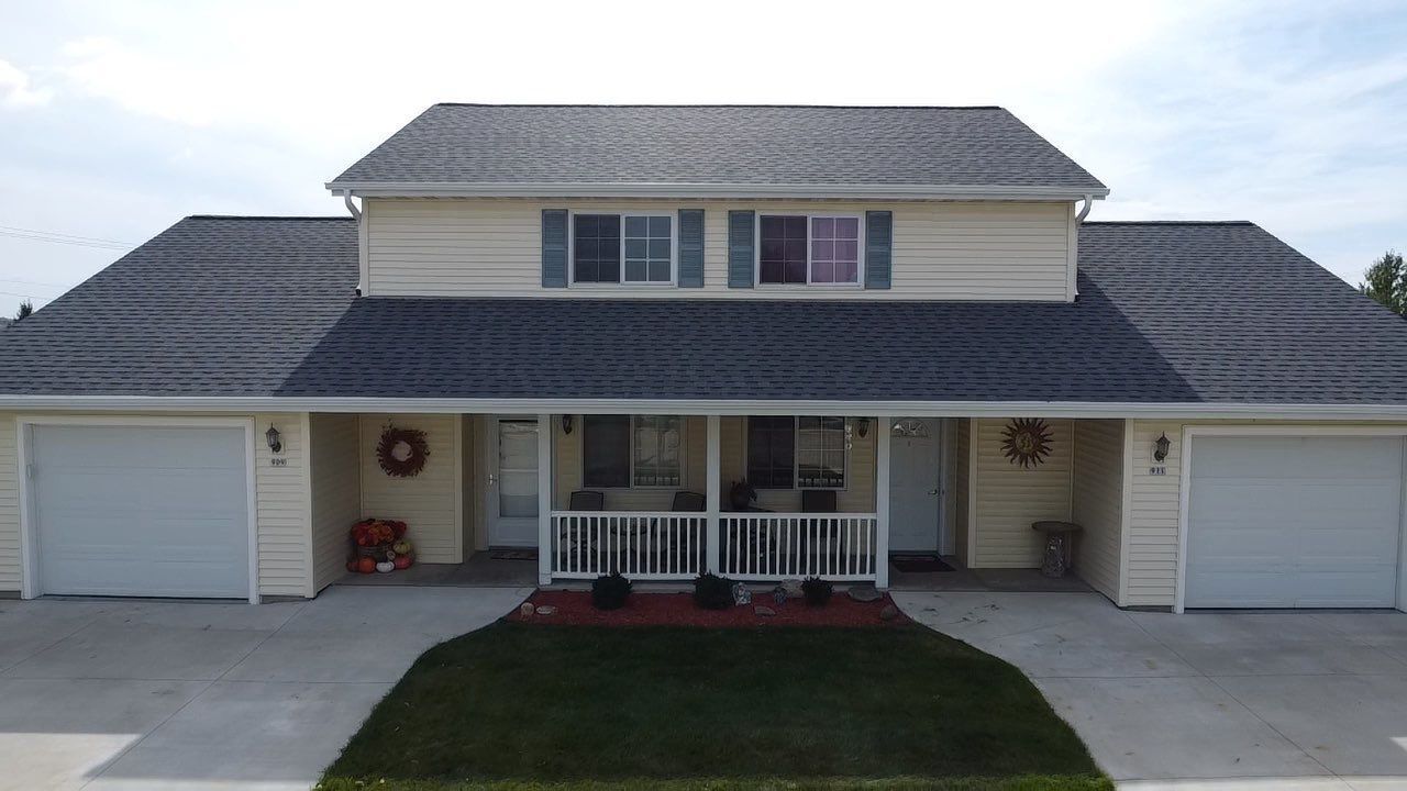 A two-story duplex with beige siding, dark gray shingled roofs, two garage doors, and a shared covered porch.