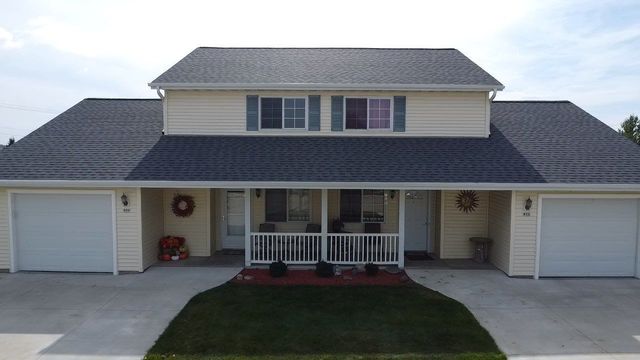A duplex home with light yellow siding, a dark roof, two garages, and a shared front porch with a white railing.