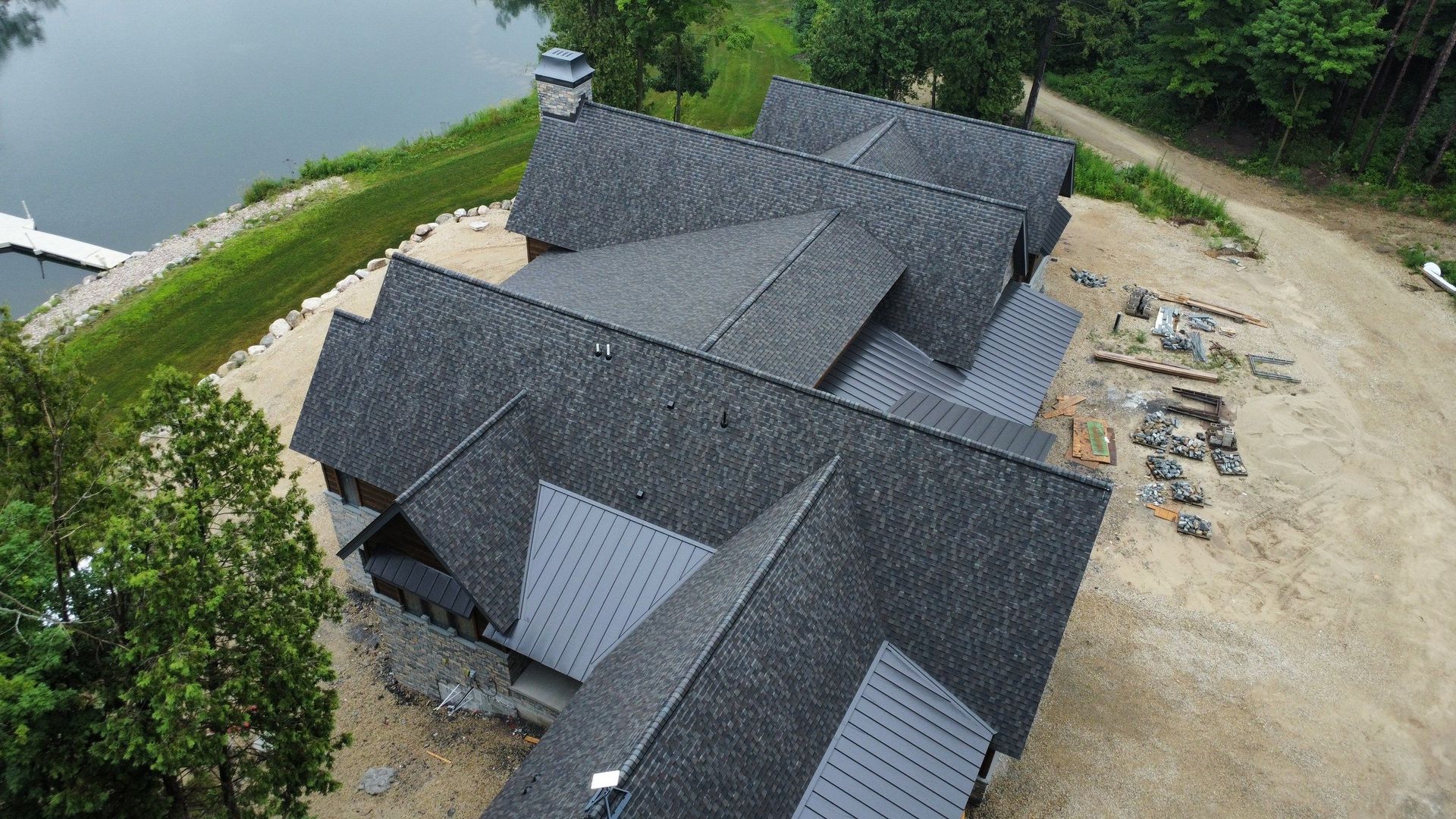 Aerial view of a house under construction with grey shingled roofs, situated near a lake and dirt grounds.
