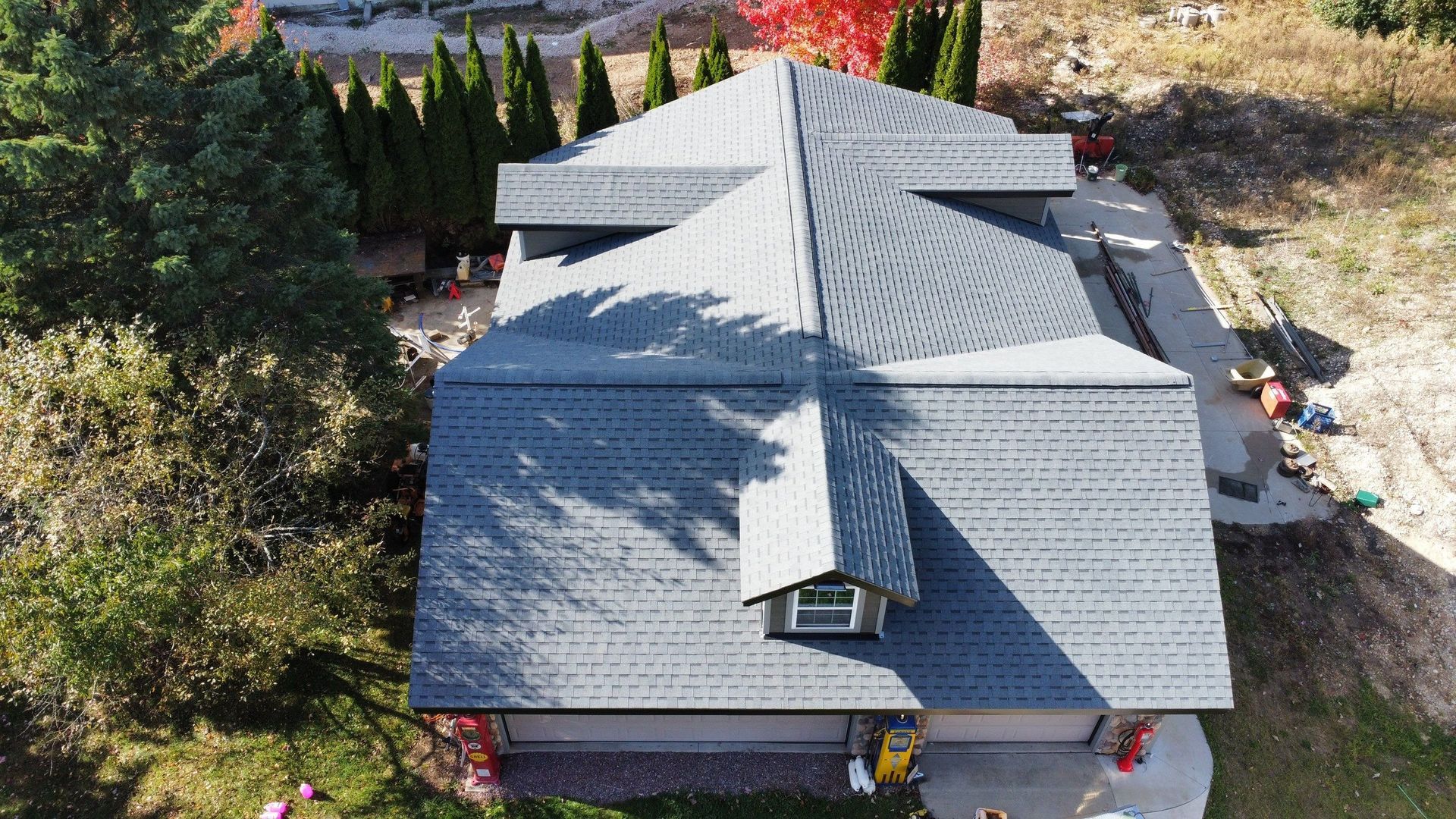 An aerial view of a gray shingled residential roof with a dormer window, surrounded by trees and a concrete driveway.