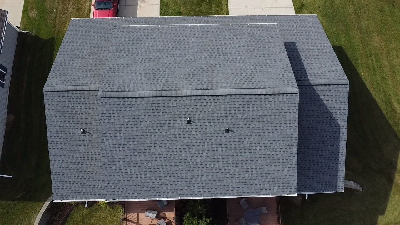 Aerial view of a gray shingled residential roof with three vent pipes and a portion of a red vehicle in the driveway.