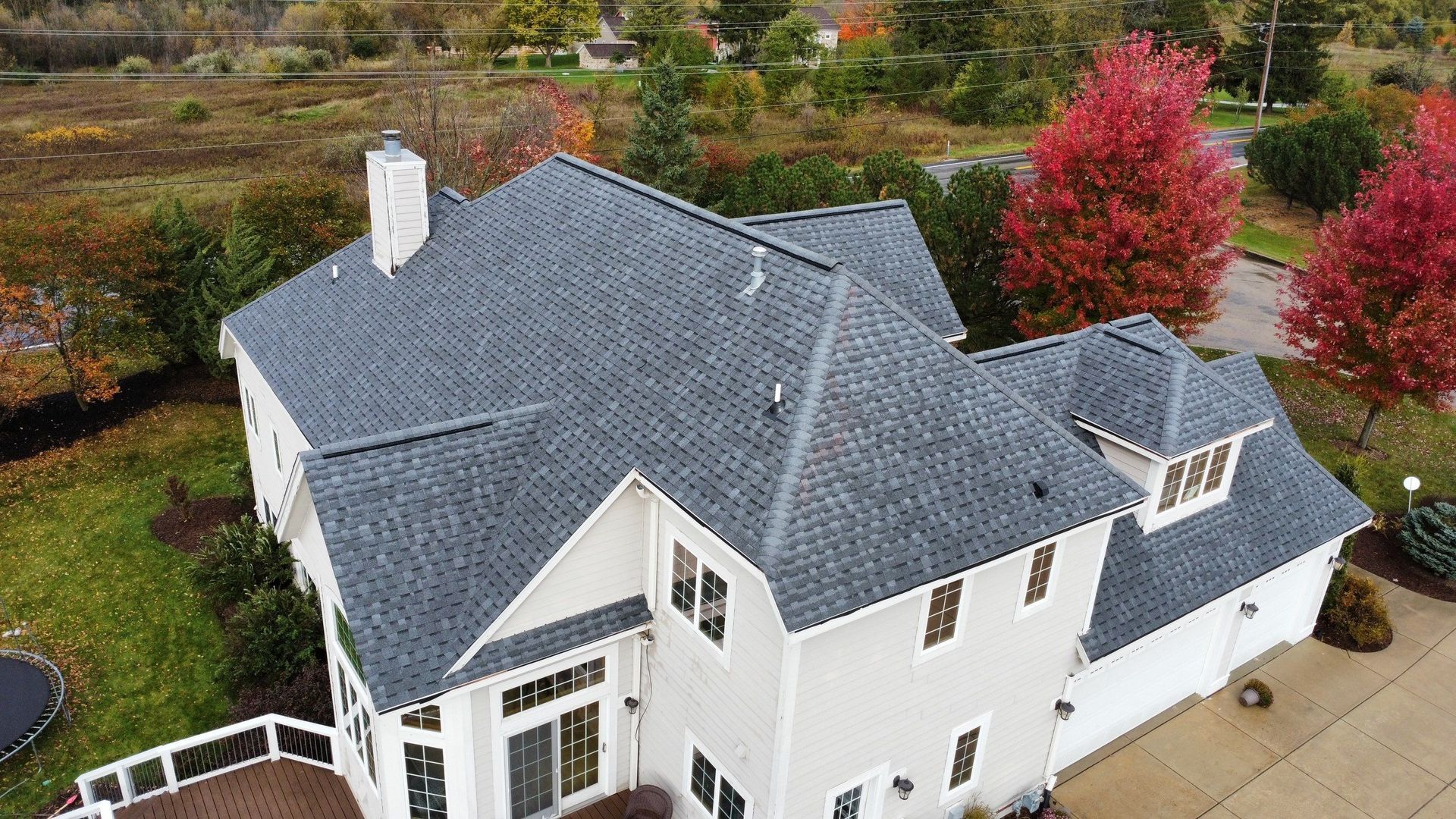 High-angle aerial view of a two-story suburban house with a dark gray shingled roof, white siding, and fall-colored trees.