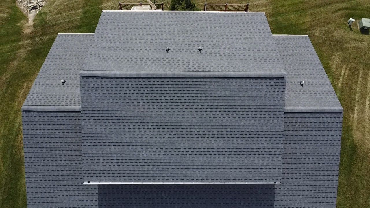 An aerial view of a dark grey shingled roof on a residential building, surrounded by a green lawn.