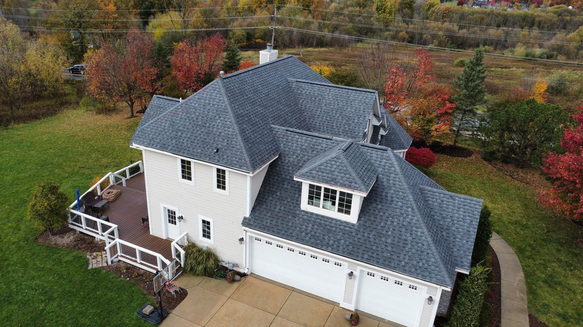 An aerial view of a two-story house with a grey shingled roof, white siding, and a wooden deck, set among autumn trees.