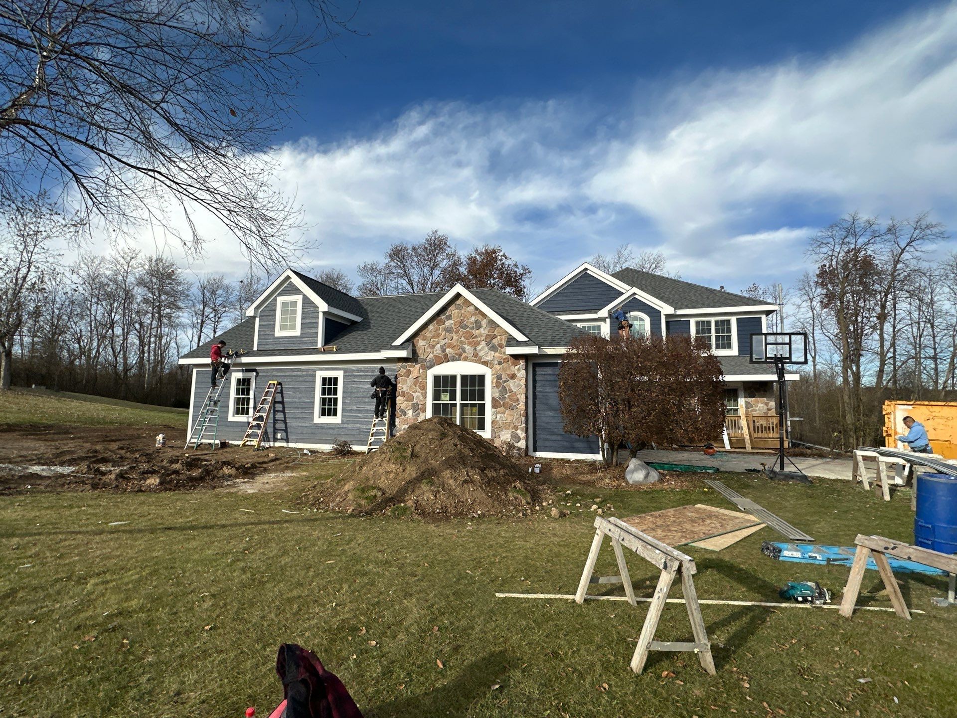 A two-story blue house with stone accents under construction, with workers on ladders and building materials in the yard.