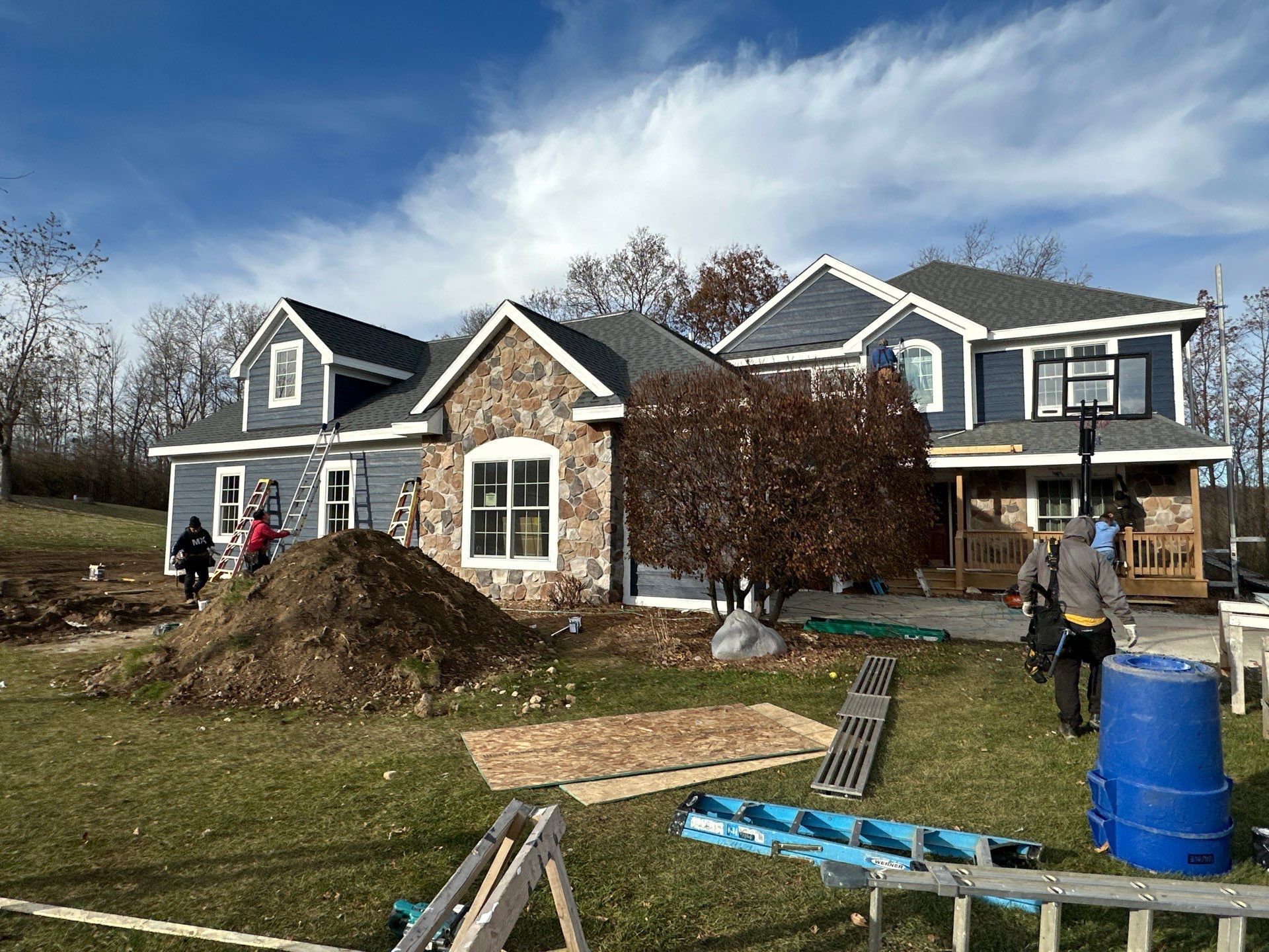 Workers at a construction site with a half-finished blue house featuring a stone facade, a large dirt pile, and equipment.