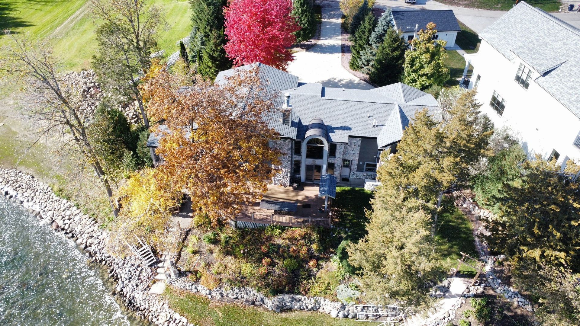 Aerial view of a large multi-story house with a grey roof, situated on a wooded shoreline next to a body of water.