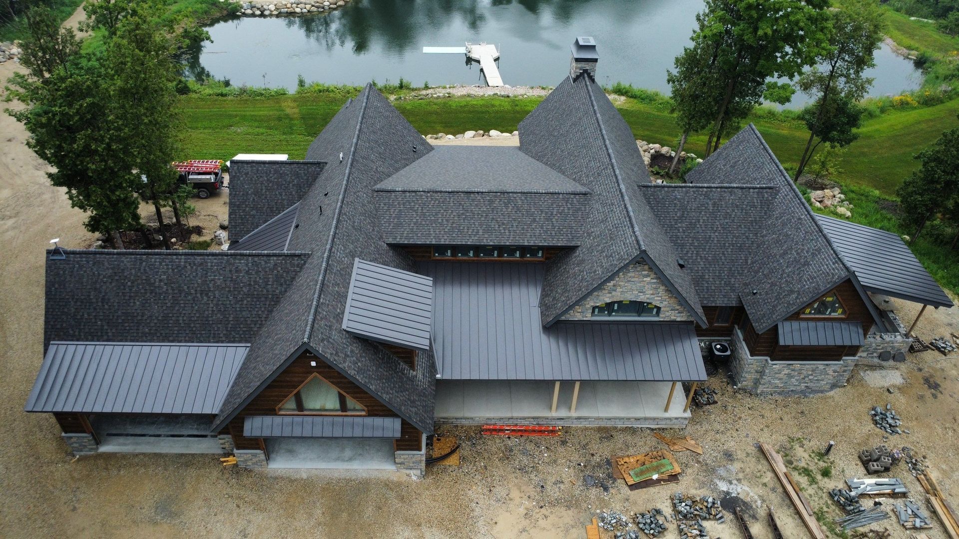 An aerial view of a large, dark-roofed luxury home under construction by a lake, featuring stone accents and a dock.