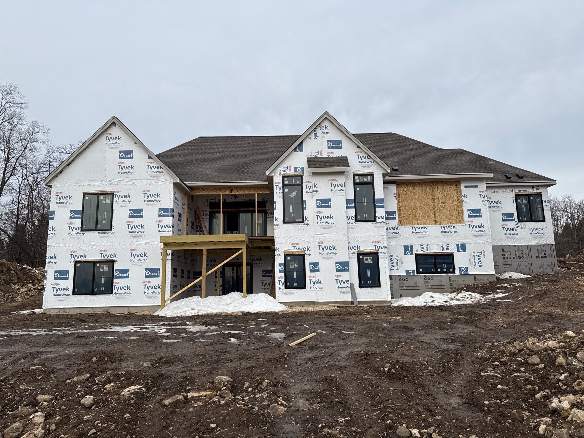 A two-story house under construction, covered in white house wrap, with a brown roof and a wooden deck structure.