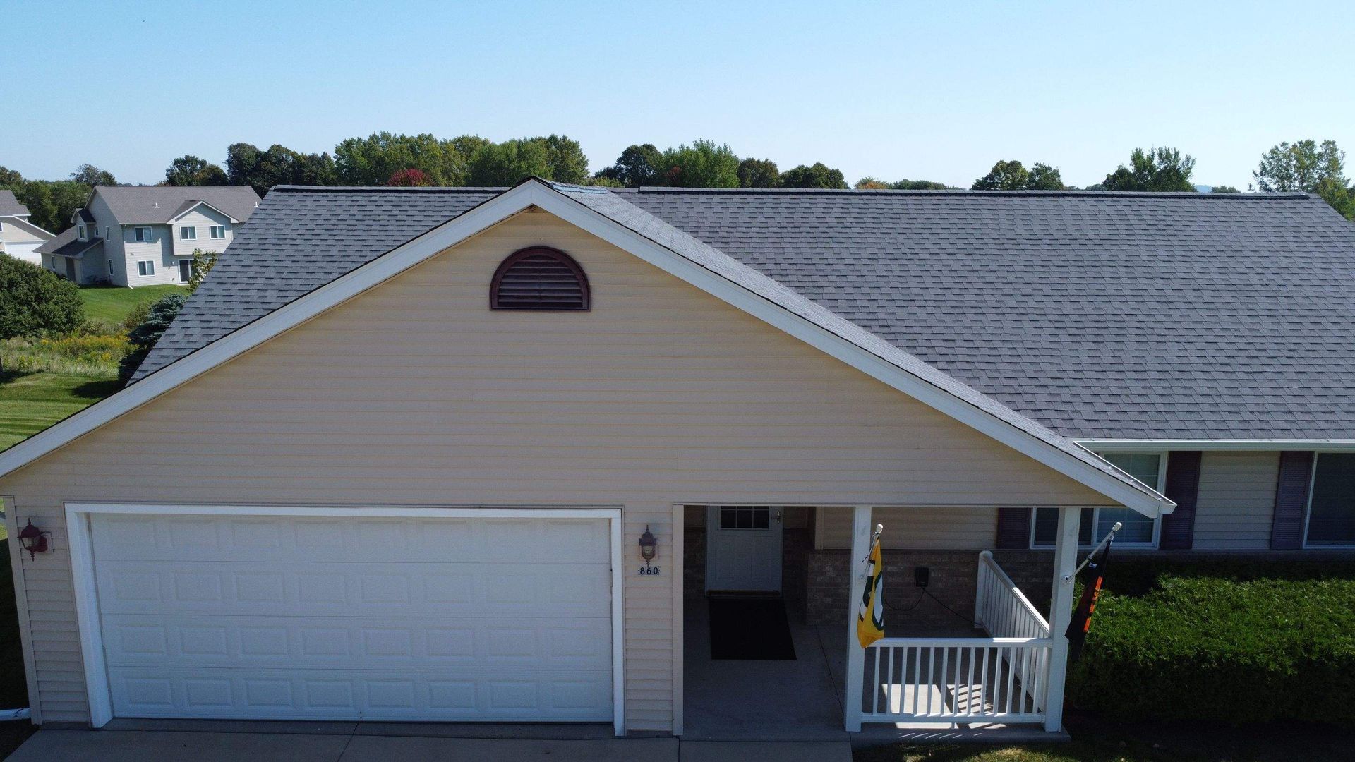 An aerial view of a tan, single-story suburban house with a gray shingled roof, a white garage door, and a front porch.