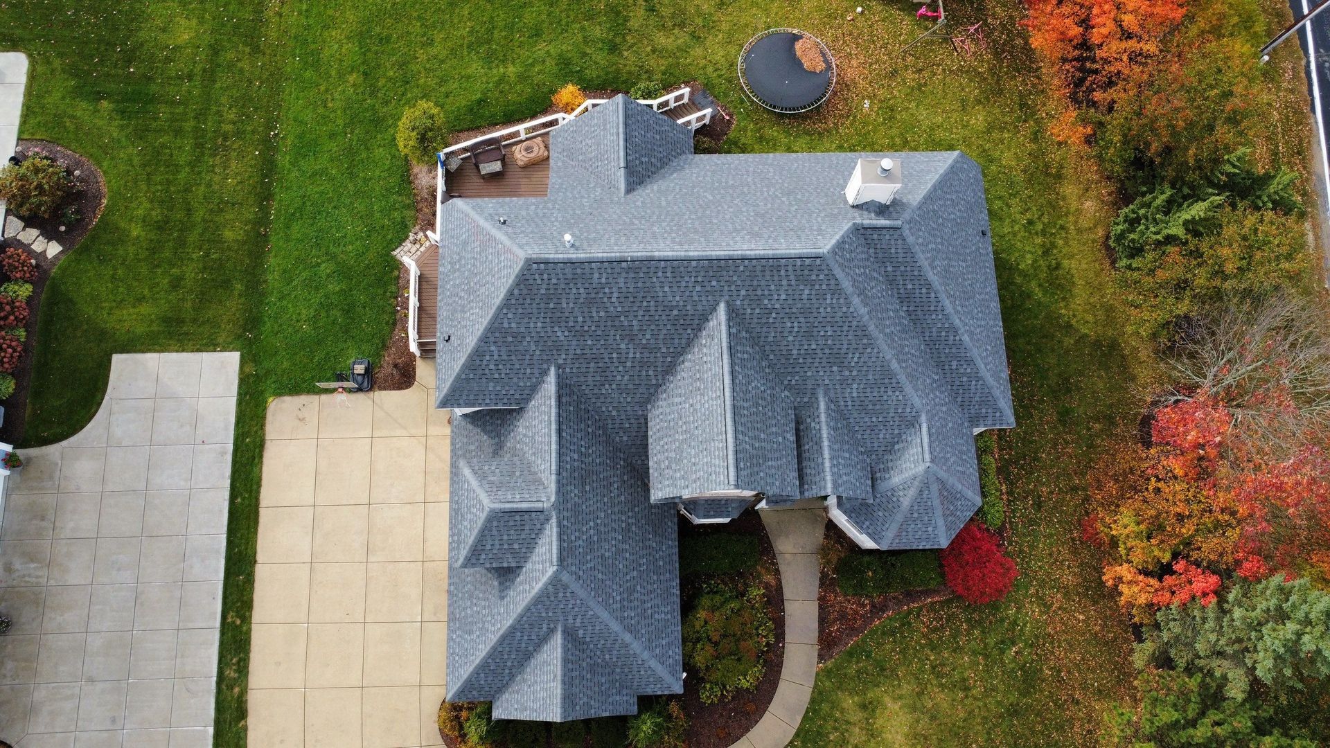 Aerial view of a house with a gray shingled roof, surrounded by a lawn, a stone patio, and autumn trees.